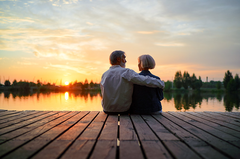 A wide, low-angle shot of a senior couple sitting on a wooden dock with their backs to the camera, looking out over a calm lake at sunset.