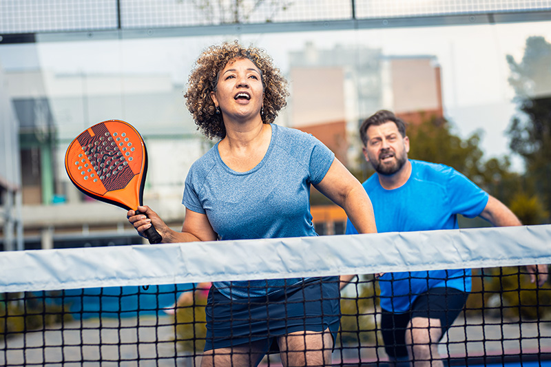 A middle-aged woman with curly brown hair and a blue shirt holds an orange paddle on a pickleball court, leaning forward over the net with a determined expression. Her partner, a man in a blue t-shirt, stands behind her.