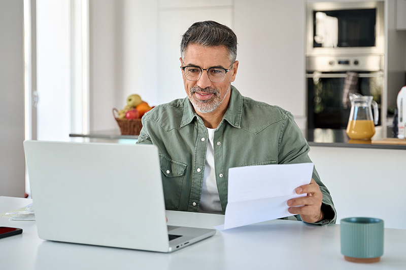 middle-aged-man-looking-at-computer-1545166937 A middle-aged man with glasses and a beard sits at a table in a bright kitchen, reviewing a paper document while looking at his open laptop.