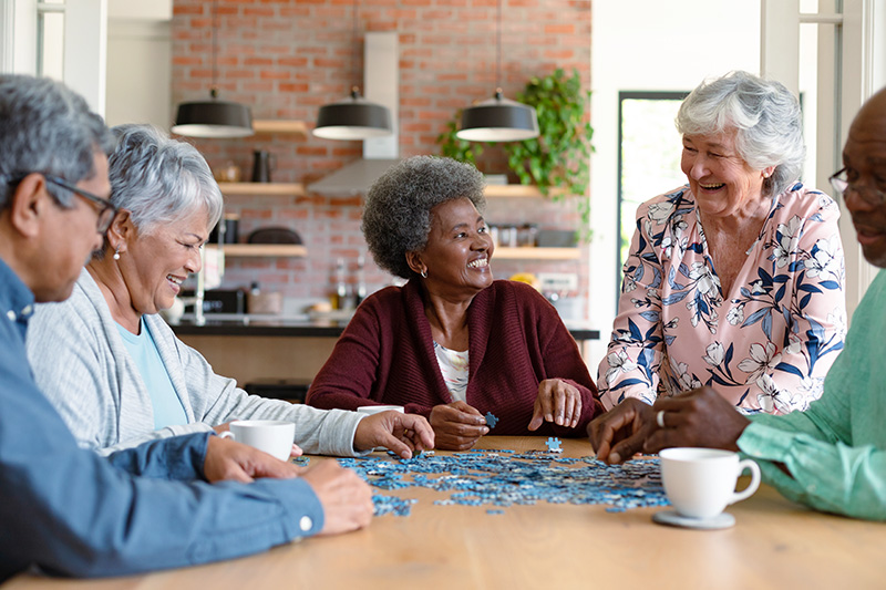 senior-friends-doing-a-puzzle_459705020 A group of five smiling seniors sitting around a wooden table, collaborating on a blue jigsaw puzzle.