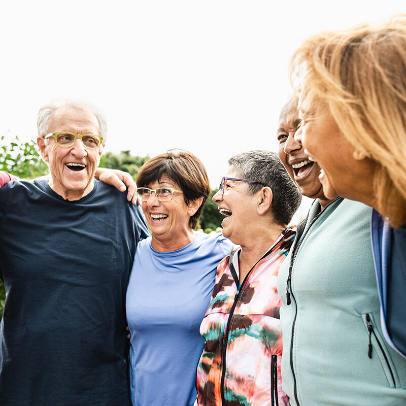 A group of five diverse seniors standing outdoors with their arms around each other, laughing heartily.