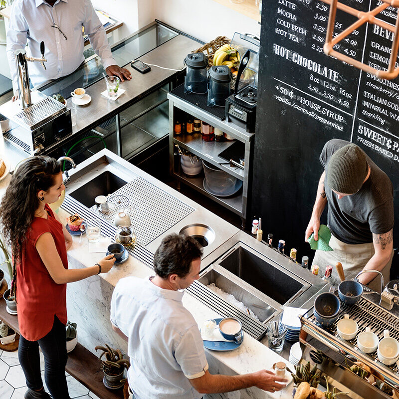 A high-angle view of a busy, modern coffee shop counter with customers ordering and baristas working behind the counter.