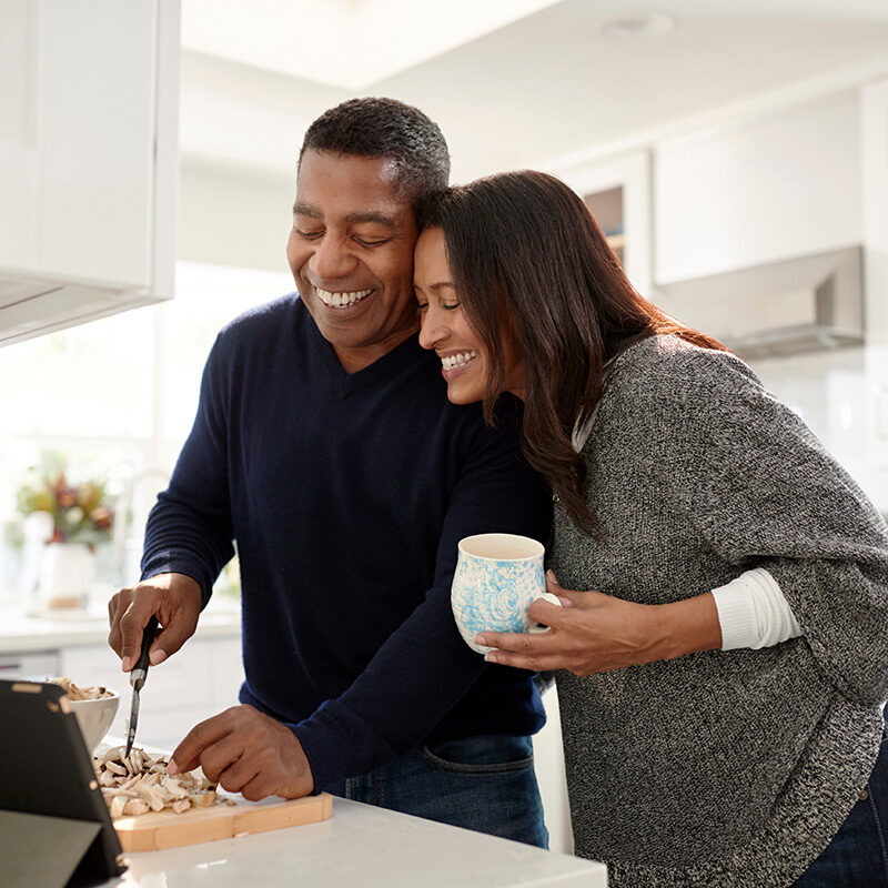 A candid, close-up photo of a smiling middle-aged, multi-racial couple cooking together in a bright, modern kitchen.