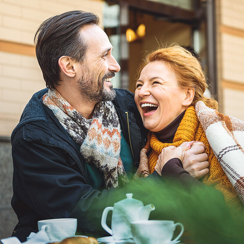 A candid, close-up outdoor photo of a happy middle-aged couple sharing a laugh while sitting at a cafe table.