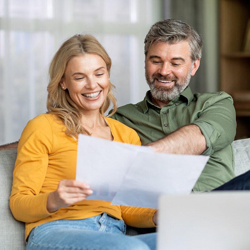 A smiling, middle-aged couple sitting on a couch, reviewing papers and looking at a laptop.
