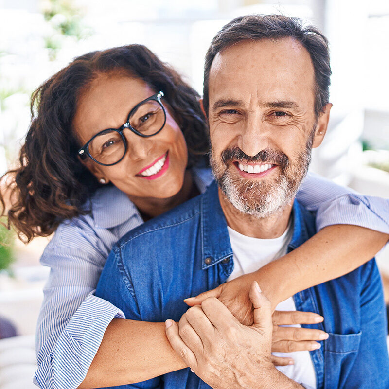 middle-aged-smiling-couple-491266631 A close-up, front-facing portrait of a smiling middle-aged, multi-racial couple. The woman, wearing glasses, is hugging the man in a denim shirt from behind.