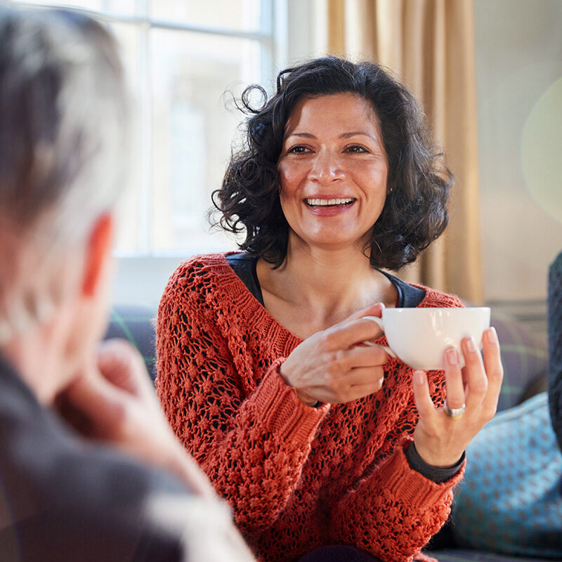 A candid, close-up photo of a smiling, middle-aged woman with dark, curly hair, wearing an orange, open-knit sweater, sitting indoors and holding a white teacup.