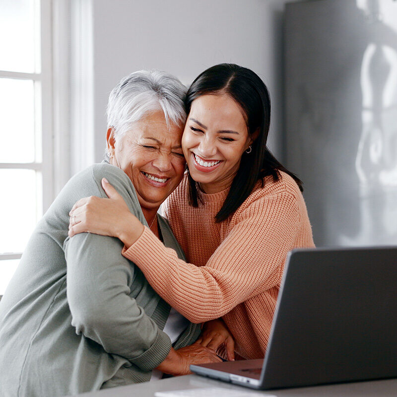 A close-up of a smiling, dark-haired younger woman in a peach-colored sweater hugging an older woman with short gray hair and a light green cardigan.