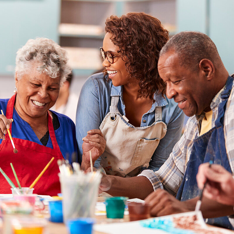 A cheerful indoor scene showing an intergenerational painting class. An African American woman in a denim shirt and apron smiles while standing between two seniors who are focused on painting.