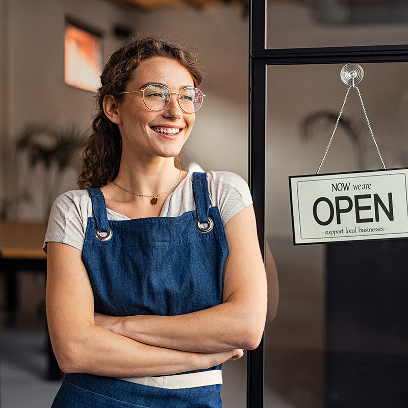 A portrait of a happy young woman, a small business owner, standing in her shop doorway with her arms crossed.